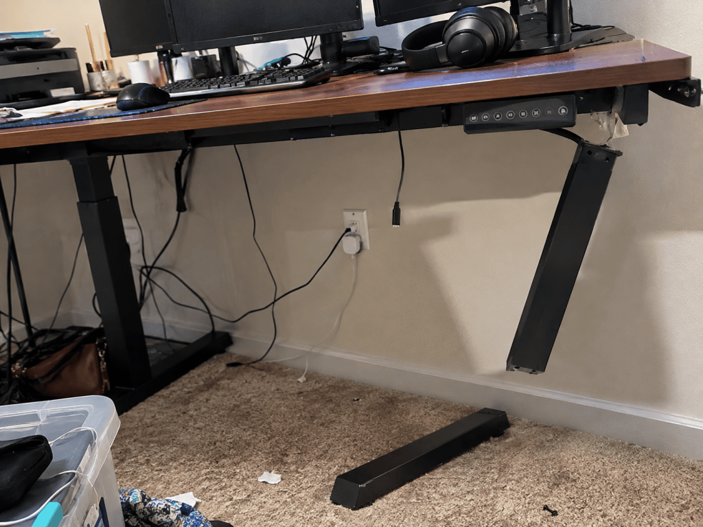 Realistic home office standing desk with one support leg detached and dangling while the desk remains loaded with equipment